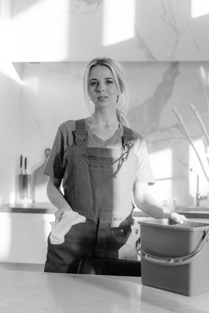 Portrait noir et blanc d’une employée de ménage tenant du matériel de nettoyage dans une cuisine moderne.